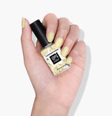 A hand with light yellow painted nails holds a bottle of LONDONTOWN Buttercup UV/LED Gel Color nail polish against a white background, showcasing the pastel yellow creme shade.