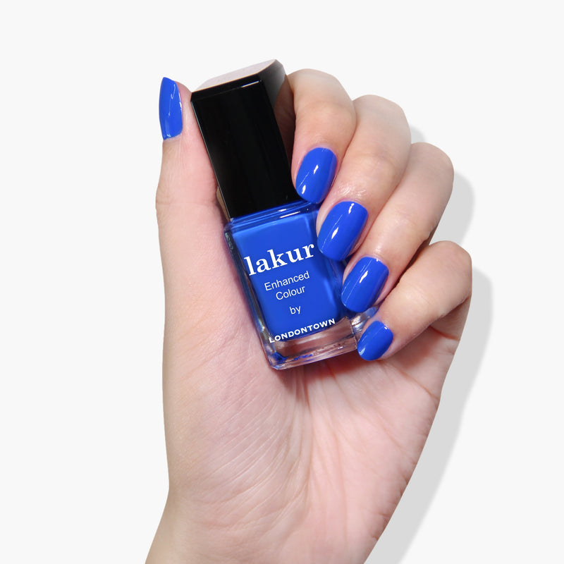 A hand with blue painted nails holds a bottle of LONDONTOWN's Iconic Lakur Enhanced Colour nail polish in matching electric blue—winner of the 2019 Allure Best of Beauty Award and proudly vegan and cruelty-free.