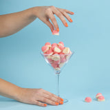 A hand with orange nails drops LONDONTOWN's Paloma, a pink and white gummy candy, into a martini glass brimming with similar treats, all set against a light blue background that captures the vibrant essence of California peach.