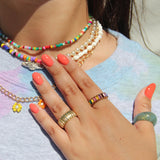 Close-up image of a person wearing colorful beaded necklaces, various gold rings, and Papaya creamy coral vegan nail polish by LONDONTOWN, with a light blue top.
