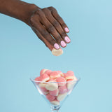 A hand adorned with LONDONTOWN's Pink Palm nails hovers over a glass filled with pink and white gummy candies, set against a blue background.